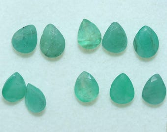 Puede incluir: Colección de nueve piedras preciosas de color verde esmeralda, cada una con forma de lágrima. Las piedras, de aspecto pulido y translúcido, varían ligeramente en tamaño y diseño de facetas. Se disponen sobre una superficie blanca.