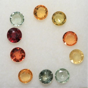 May include: Assortment of nine round gemstones in various colors, including red, orange, yellow, and green. The gemstones are arranged in a circle on a white surface.