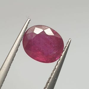 Puede incluir: Una gema ovalada de color rojo vibrante con un corte facetado. La gema se sostiene con unas pinzas sobre un fondo blanco.