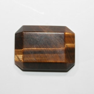 May include: A rectangular tiger's eye gemstone with a polished, faceted surface. The stone displays bands of brown and gold, with a dark brown base. The gemstone has an emerald cut with beveled edges.