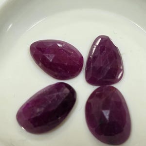 Puede incluir: Cuatro gemas facetadas, de forma ovalada y de color rojo intenso. Las gemas están dispuestas en un patrón circular sobre un fondo blanco. Las piedras tienen una superficie pulida y reflectante.