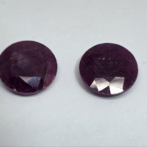 Puede incluir: Dos gemas redondas, facetadas, de color rojo intenso. Las gemas tienen una superficie pulida con estructuras internas visibles y facetas reflectantes. Las piedras se colocan sobre un fondo blanco, resaltando su color y forma.
