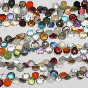 Könnte beinhalten: Eine Sammlung facettierter Edelsteine in verschiedenen Farben, darunter Rot, Orange, Gelb, Grün, Blau, Lila und Klar. Die Edelsteine sind auf einer weißen Oberfläche angeordnet und zeigen ihre unterschiedlichen Formen und Größen. Ideal für die Schmuckherstellung.