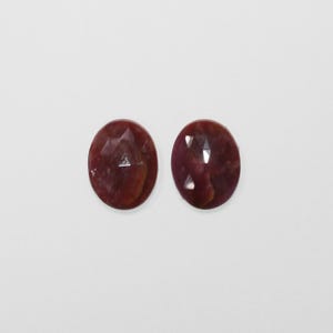 Könnte beinhalten: Zwei ovale, facettierte Edelsteine in tiefroter Farbe. Die Edelsteine haben eine polierte Oberfläche mit sichtbaren inneren Strukturen und sind vor einem schlichten weißen Hintergrund platziert. Geeignet für Schmuckherstellung.