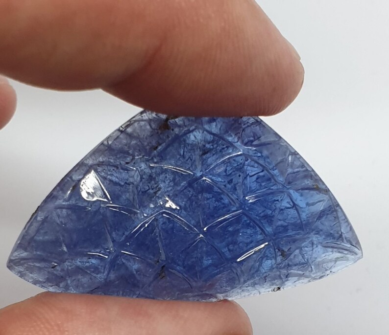 May include: A carved blue gemstone with a geometric pattern. The gemstone is shaped like a triangle and has a smooth, polished surface.
