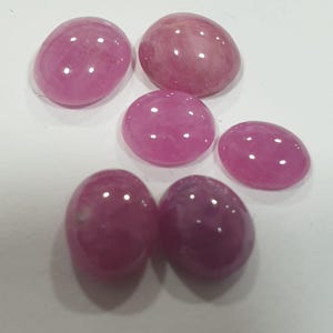Könnte beinhalten: Eine Sammlung von sechs polierten, oval geformten Edelsteinen in verschiedenen Rosa- und Magentatönen. Die Steine haben eine glatte, glänzende Oberfläche, die das Licht reflektiert. Die Edelsteine sind auf einer weißen Oberfläche angeordnet.