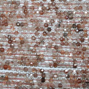 May include: Close-up of a strand of faceted gemstone beads. The beads are a mix of clear, brown, and reddish-brown hues, strung together with thin, light red string. The beads have a square or rectangular shape.