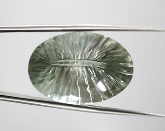 May include: An oval-shaped, faceted gemstone in shades of green is held by a pair of silver tweezers. The gemstone has a detailed, radial pattern of cuts, creating a textured surface that reflects light. The gem is set against a plain white background.