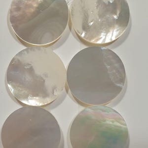 Könnte beinhalten: Sechs runde, schillernde Perlmuttmuschelteile. Die Muscheln haben einen perlmuttartigen Glanz mit Farben von Weiß bis Hellrosa und Grün. Die kreisförmigen Formen sind in zwei Reihen zu je drei Stück auf weißem Hintergrund angeordnet.