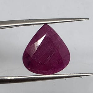 May include: A pear-shaped, faceted gemstone with a deep magenta-red color. The stone features a smooth, polished surface with visible internal inclusions and geometric facets that reflect light, held securely by a pair of metal tweezers.