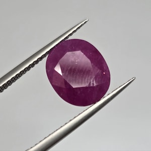 Puede incluir: Una gema de rubí facetada, de forma ovalada, sujeta con pinzas. El rubí es de un color rojo intenso y rico con una superficie pulida. Las pinzas son plateadas y el fondo es gris neutro.