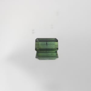 May include: A faceted, rectangular green gemstone with a reflective surface. The gem has a dark green hue and appears to be transparent. The gemstone is likely a tourmaline or similar precious stone, suitable for jewelry making.
