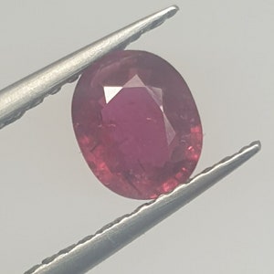 May include: A vibrant, oval-shaped ruby gemstone with a deep red hue. The gemstone is held in place by a pair of tweezers.