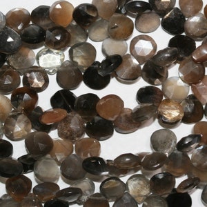 May include: A collection of faceted heart-shaped beads in various shades of brown, grey, and black. Each bead features a polished surface with geometric facets that reflect light, showcasing natural colour variations and internal inclusions.