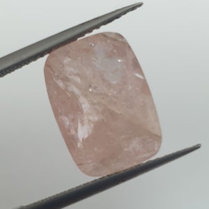 Pode incluir: Uma gema rosa pálida com formato retangular, segurada por uma pinça. A gema tem uma aparência translúcida com inclusões internas visíveis.