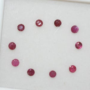 Könnte beinhalten: Eine Ansammlung von elf kleinen, runden Rubin-Edelsteinen. Die Edelsteine haben eine tiefrote Farbe und scheinen facettiert zu sein. Sie sind in einem kreisförmigen Muster auf weißem Hintergrund angeordnet.