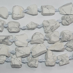 Può includere: Una collana di perle di howlite bianche, ogni perla ha una forma naturale irregolare e una superficie ruvida e testurizzata. Le perle sono infilate su un cordoncino bianco con nodi tra ogni perla.