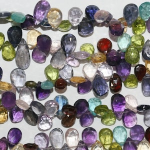 Puede incluir: Una colección de gemas facetadas en varios colores, incluyendo púrpura, verde, azul y transparente. Las gemas tienen forma de lágrima y redonda, ensartadas juntas. La imagen es un primer plano, mostrando los diferentes colores y cortes de las piedras.
