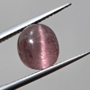 May include: A polished, oval-shaped gemstone in a soft mauve hue, held by silver tweezers. The gem has a chatoyant effect, with a band of light across its surface. The background is a neutral gray.