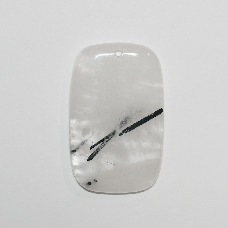 May include: A white quartz pendant with black tourmaline inclusions. The pendant is shaped like a rectangle with rounded edges and has a hole at the top for hanging.