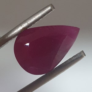 Peut inclure: Une pierre précieuse rose foncé en forme de poire, tenue par des pinces. La pierre précieuse est facetée et a un haut poli.