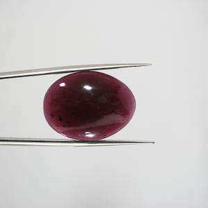 Könnte beinhalten: Ein polierter, oval geformter, tiefroter Edelstein wird von einer silbernen Pinzette vor einem weißen Hintergrund gehalten. Der Edelstein hat eine glatte, glänzende Oberfläche mit subtilen Farbvariationen.