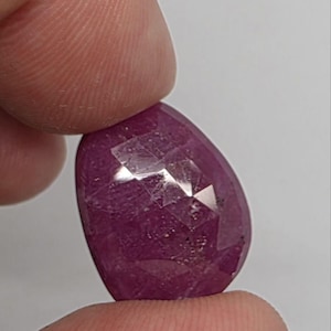 May include: A faceted, oval-shaped ruby gemstone. The gem is a deep, rich red color with multiple facets that reflect light. The ruby is held between two fingers against a plain white background.