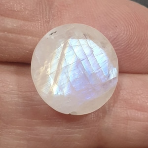 May include: A round, faceted moonstone gemstone. The stone is mostly white with iridescent blue and purple flashes. The gemstone is held in a hand, showing the natural variations and beauty of the stone.