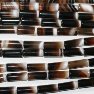 May include: Close-up of multiple strands of cylindrical beads. The beads are a mix of black, brown, and white, with a polished, glossy finish. The beads are arranged in rows, showcasing their varied color patterns and shapes.
