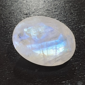 May include: Oval-shaped, polished moonstone gemstone. The stone has a milky white base with iridescent blue and purple flashes. The surface is smooth and reflective, with internal fractures visible. The gemstone is set against a dark background.
