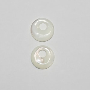 May include: Two white mother of pearl shell rings with a hole in the center. The rings are smooth and have a slight iridescent sheen.