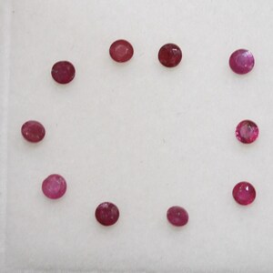 May include: Ten small, round ruby gemstones in varying shades of red and pinkish-red. The gems are displayed on a white surface, highlighting their polished appearance. The gemstones are arranged in a circular pattern.