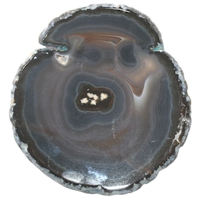 Peut inclure: Une tranche de pierre d'agate polie avec des bandes concentriques de gris, de brun et de blanc. La pierre a un bord extérieur gris foncé et une zone centrale avec de petits amas de cristaux blancs. La forme générale est approximativement circulaire.