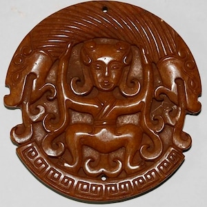 May include: A brown jade pendant with a carved image of a mythical figure with a crown and a decorative border.