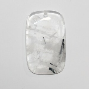 May include: A clear quartz pendant with black tourmaline inclusions. The pendant is rectangular in shape with rounded edges and has a hole drilled at the top for hanging.