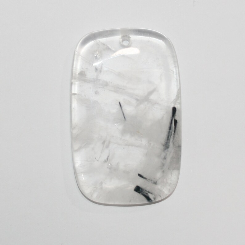 May include: A clear quartz pendant with black tourmaline inclusions. The pendant is rectangular in shape with rounded edges and has a hole drilled at the top for hanging.