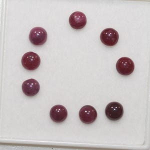 Peut inclure: Huit rubis ronds taillés en cabochon disposés en cercle. Chaque pierre précieuse présente une teinte rouge profonde et saturée avec une surface lisse et polie. Les pierres sont présentées sur un plateau de présentation blanc uni.