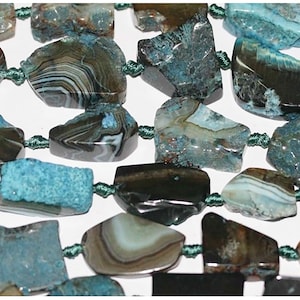 Puede incluir: Una hebra de cuentas de piedra de ágata azul y negra. Las cuentas son de forma irregular y tienen una textura natural y rugosa. Las cuentas están enhebradas en un cordón verde oscuro.