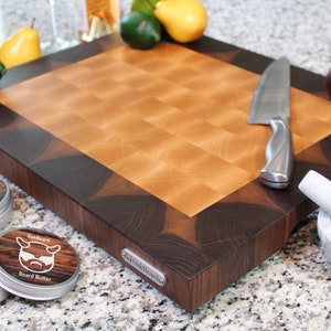 May include: A square wooden cutting board with a light-colored checkered center and a dark wood border. A metal badge labeled OakBeard is on the side. Three round tins of OakBeard Board Butter, a silver knife, and a white marble mortar and pestle.
