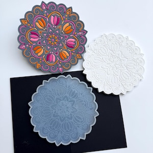 Può includere: Tre sottobicchieri rotondi con disegni mandala. Uno è grigio con dettagli rosa, arancioni e gialli. Un altro è bianco con un motivo in rilievo. Il terzo è un blu-grigio traslucido. Tutti hanno bordi smerlati.