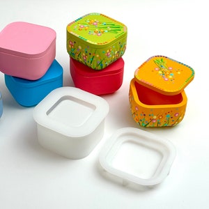 Peut inclure: Cinq boîtes carrées avec des couvercles. Les boîtes sont en bois peint et ont des couleurs et des motifs différents. Les boîtes sont empilées les unes sur les autres. Deux des boîtes sont ouvertes, révélant l'intérieur des boîtes. Les boîtes sont sur une surface blanche.