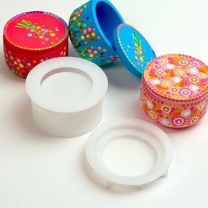 May include: Three round, painted, decorative boxes with lids. The boxes are painted with colorful dots and swirls. Two of the boxes are open and the lids are beside them. Two white silicone molds are also shown. The molds are for making the boxes.