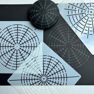 Op de afbeelding: Drie witte plastic sjablonen met ronde ontwerpen. De sjablonen liggen op een zwart oppervlak met een zwarte bol met een wit rasterpatroon.