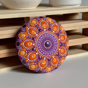 Handbemalter Mandala-Stein als dekoratives Kunstobjekt auf einer Holzablage im Wohnbereich. Das Dot Painting in Orange, Apricot, Lila und Flieder hebt sich kontrastreich von der magentafarbenen Grundierung ab. Die symmetrische Mandala-Gestaltung mit ovalen und runden Punktformen verleiht dem Unikat eine harmonische Ausstrahlung und macht es zu einem besonderen Blickfang für Wohnräume und kreative Rückzugsorte.