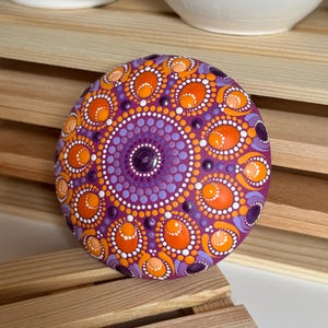 Detailansicht eines handbemalten Mandala-Steins mit magentafarbener Grundierung und Dot Painting in Orange, Apricot, Lila und Flieder. Die Aufnahme zeigt die feinen Übergänge zwischen den Farben sowie die Kombination aus runden und ovalen Dot-Elementen. Das symmetrische Mandala wirkt durch die erhabenen Farbpunkte besonders plastisch und hebt sich dekorativ von der natürlichen Holzstruktur im Hintergrund ab.