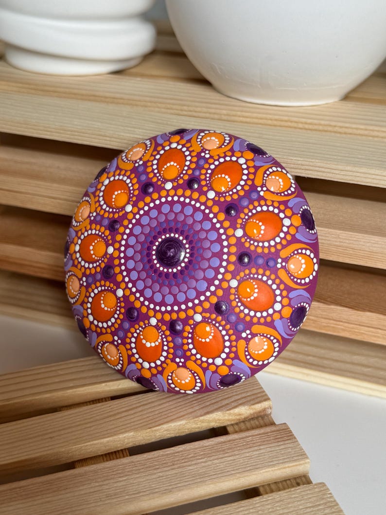 Detailansicht eines handbemalten Mandala-Steins mit magentafarbener Grundierung und Dot Painting in Orange, Apricot, Lila und Flieder. Die Aufnahme zeigt die feinen Übergänge zwischen den Farben sowie die Kombination aus runden und ovalen Dot-Elementen. Das symmetrische Mandala wirkt durch die erhabenen Farbpunkte besonders plastisch und hebt sich dekorativ von der natürlichen Holzstruktur im Hintergrund ab.
