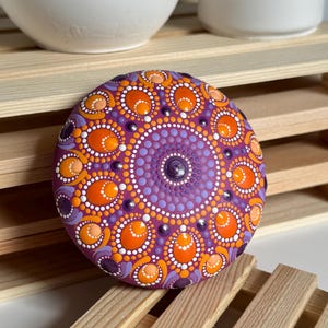 Schrägansicht eines handbemalten Mandala-Steins mit Dot Painting in Orange, Apricot, Lila und Flieder. Das symmetrische Mandala auf magentafarbenem Untergrund zeigt die besondere Wirkung der ovalen und runden Punktformen aus leicht versetzter Perspektive. Die Aufnahme hebt die Tiefe des Musters, die erhabenen Farbpunkte und die handgefertigten Details des Kunstwerks hervor.