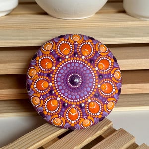 Handbemalter Mandala-Stein auf magentafarbenem Untergrund mit detailreichem Dot Painting in Orange, Apricot, Lila und Flieder. Das symmetrische Mandala zeigt runde und ovale Punktformen, die dem Kunstwerk eine besondere Tiefe verleihen. Die erhabenen Farbpunkte und die harmonische Farbgestaltung machen den Stein zu einem dekorativen Kunstobjekt für Wohnräume, Meditationsecken und kreative Bereiche.