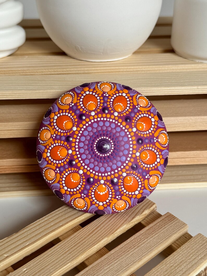 Handbemalter Mandala-Stein auf magentafarbenem Untergrund mit detailreichem Dot Painting in Orange, Apricot, Lila und Flieder. Das symmetrische Mandala zeigt runde und ovale Punktformen, die dem Kunstwerk eine besondere Tiefe verleihen. Die erhabenen Farbpunkte und die harmonische Farbgestaltung machen den Stein zu einem dekorativen Kunstobjekt für Wohnräume, Meditationsecken und kreative Bereiche.