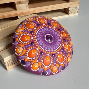 Schrägansicht eines handbemalten Mandala-Steins mit magentafarbener Grundierung und Dot Painting in Orange, Apricot, Lila und Flieder. Die Perspektive macht die gewölbte Form des Steins sowie die erhabenen Farbpunkte besonders gut sichtbar. Runde und ovale Dot-Elemente bilden ein detailreiches Mandala, das durch seine handbemalte Struktur und die harmonische Farbgestaltung eine besondere Tiefe erhält.